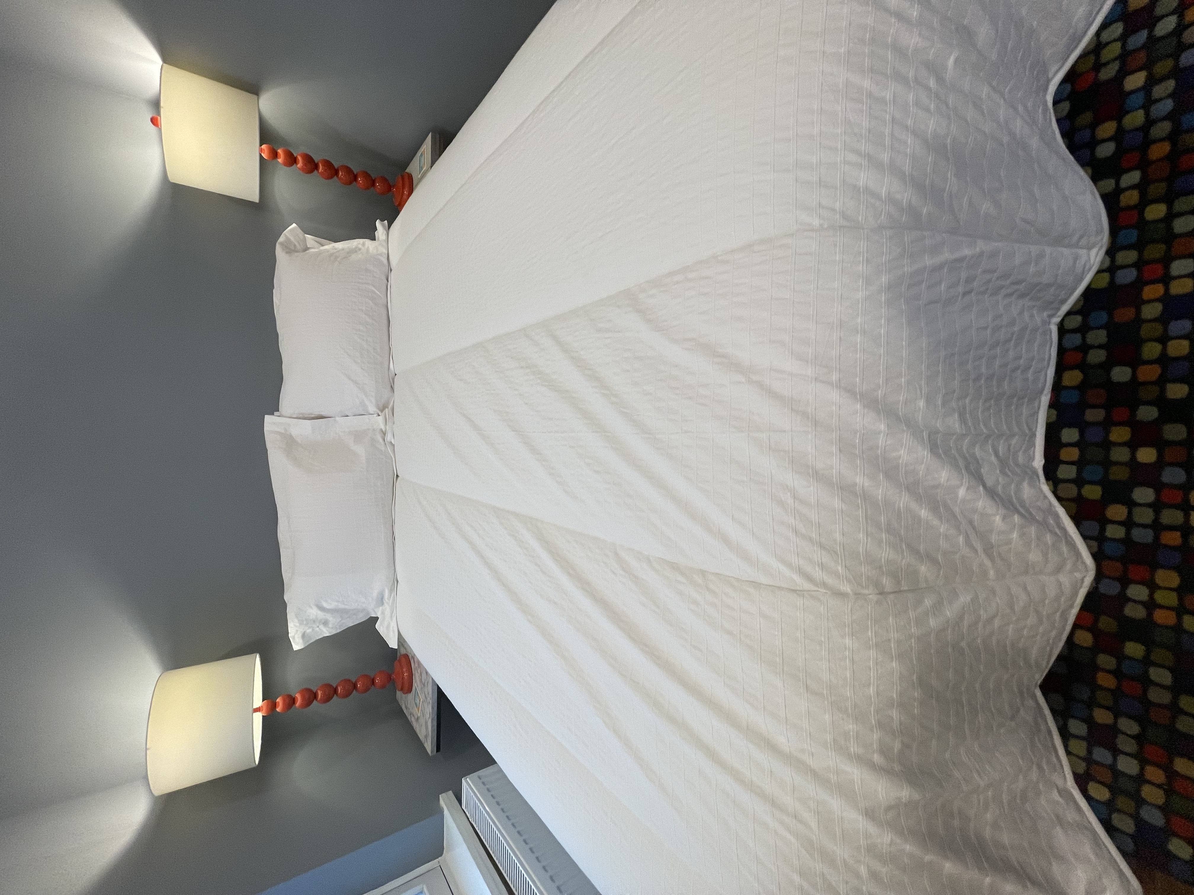 A white quilted comforter is centered on a bed with two white pillows, bedside tables with lamps on either side, and a colorful carpet in the background.