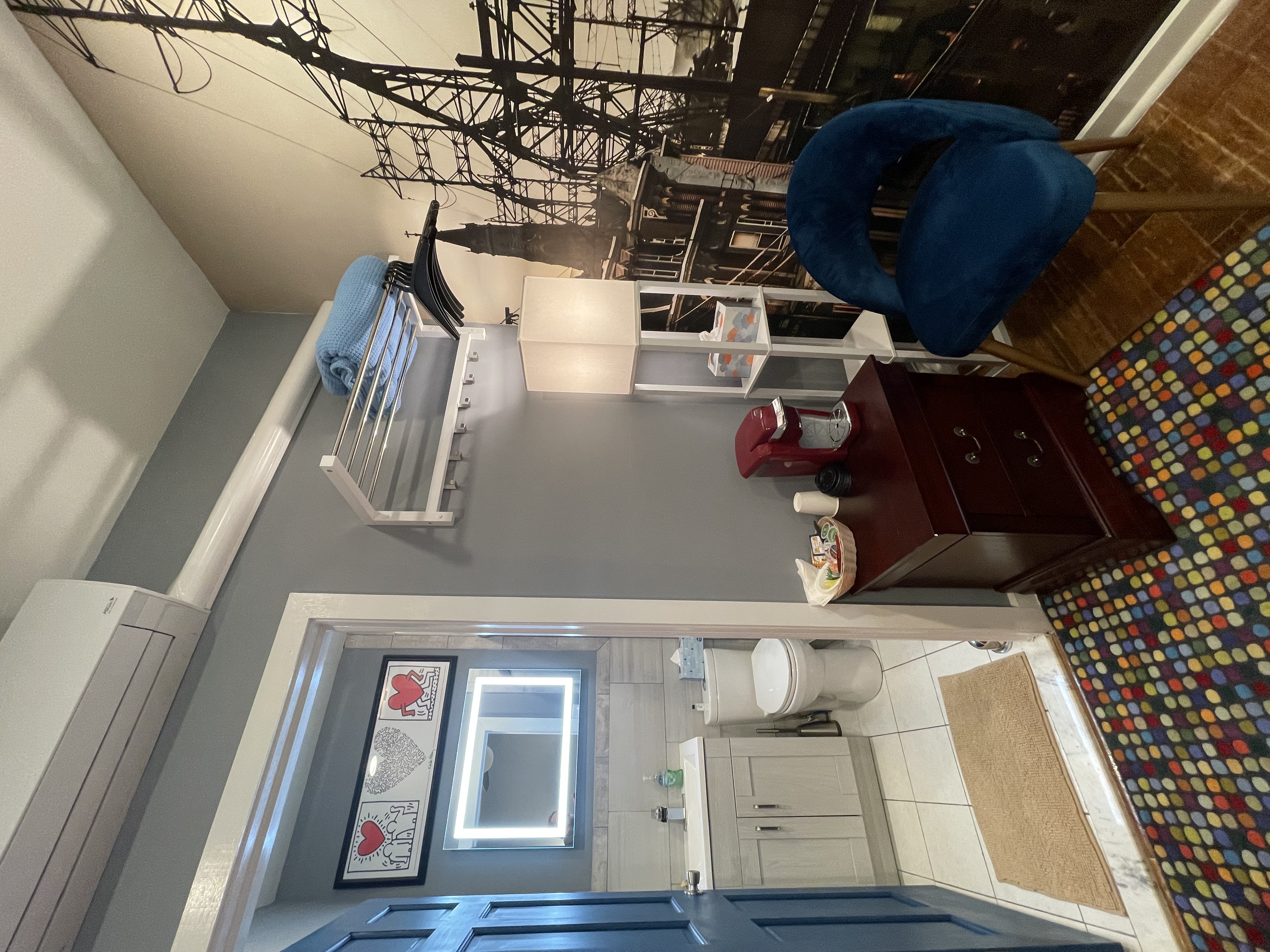 A compact entryway with a dark wood cabinet, a blue velvet chair, and a glimpse into a bathroom through an open door. A black and white mural is on the right wall.
