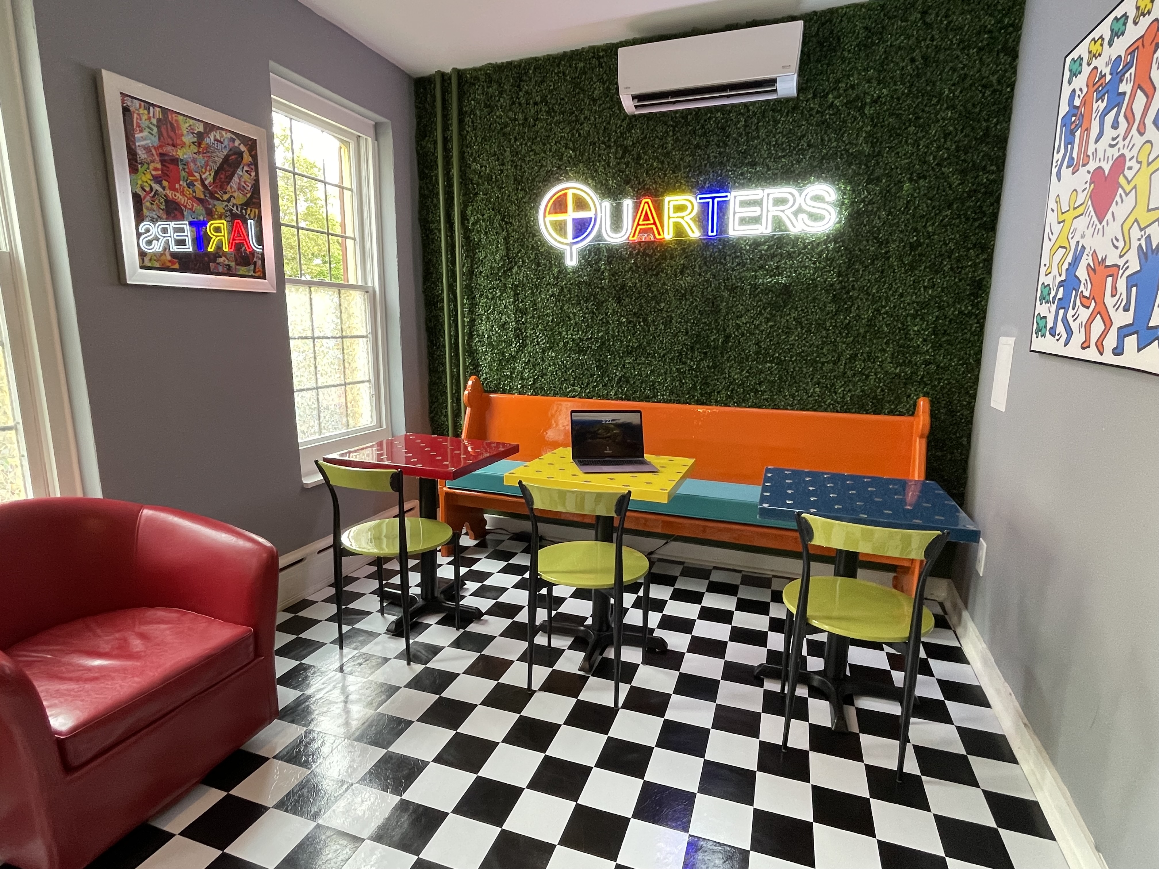 A dining area with a "Quarters" neon sign on a green textured wall, featuring several tables, a red sofa, and artwork on the walls.