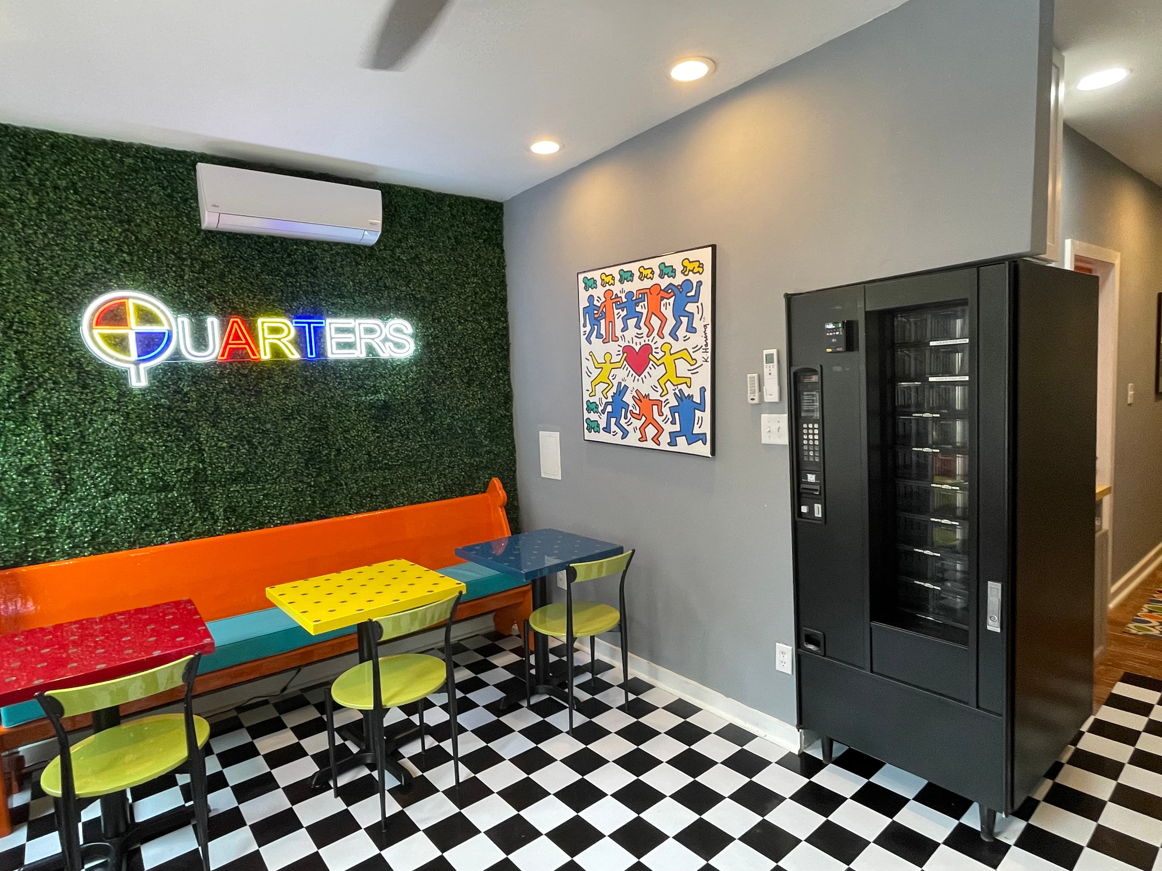 A dining area with black and white checkered floors, colorful tables and chairs, a vending machine, and a "Quarters" neon sign on a textured green wall.