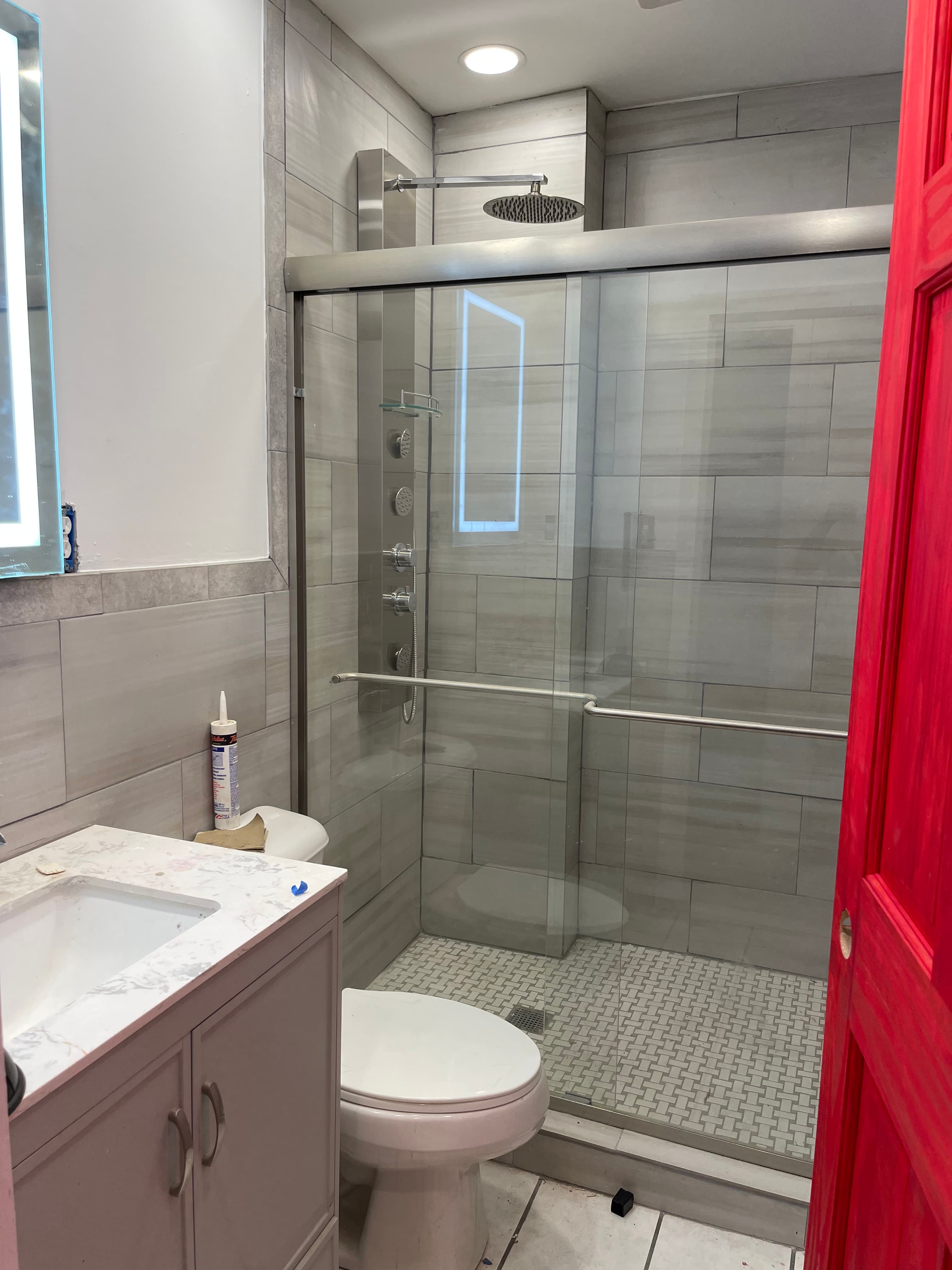 A modern bathroom with a glass-enclosed shower, a toilet, and a vanity with a sink. The walls are tiled with large, light-colored tiles. A red door is partially visible on the right side of the image.