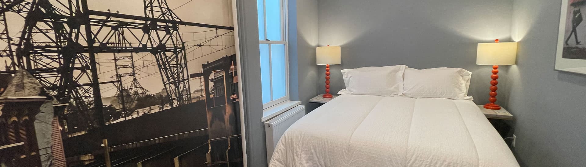 A cozy bedroom featuring a white bed, orange lamps, and a wall mural of a power line.