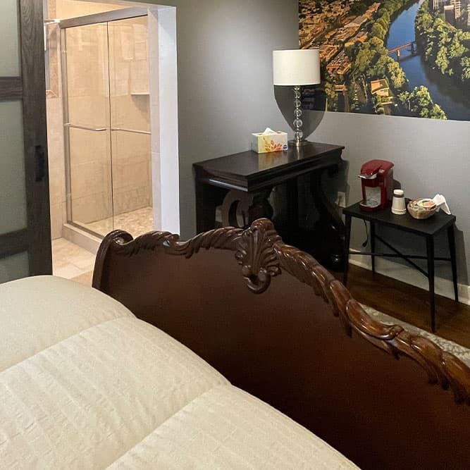 A cozy bedroom featuring a wooden bed, stylish lamp, small table with coffee setup, and an adjoining bathroom.