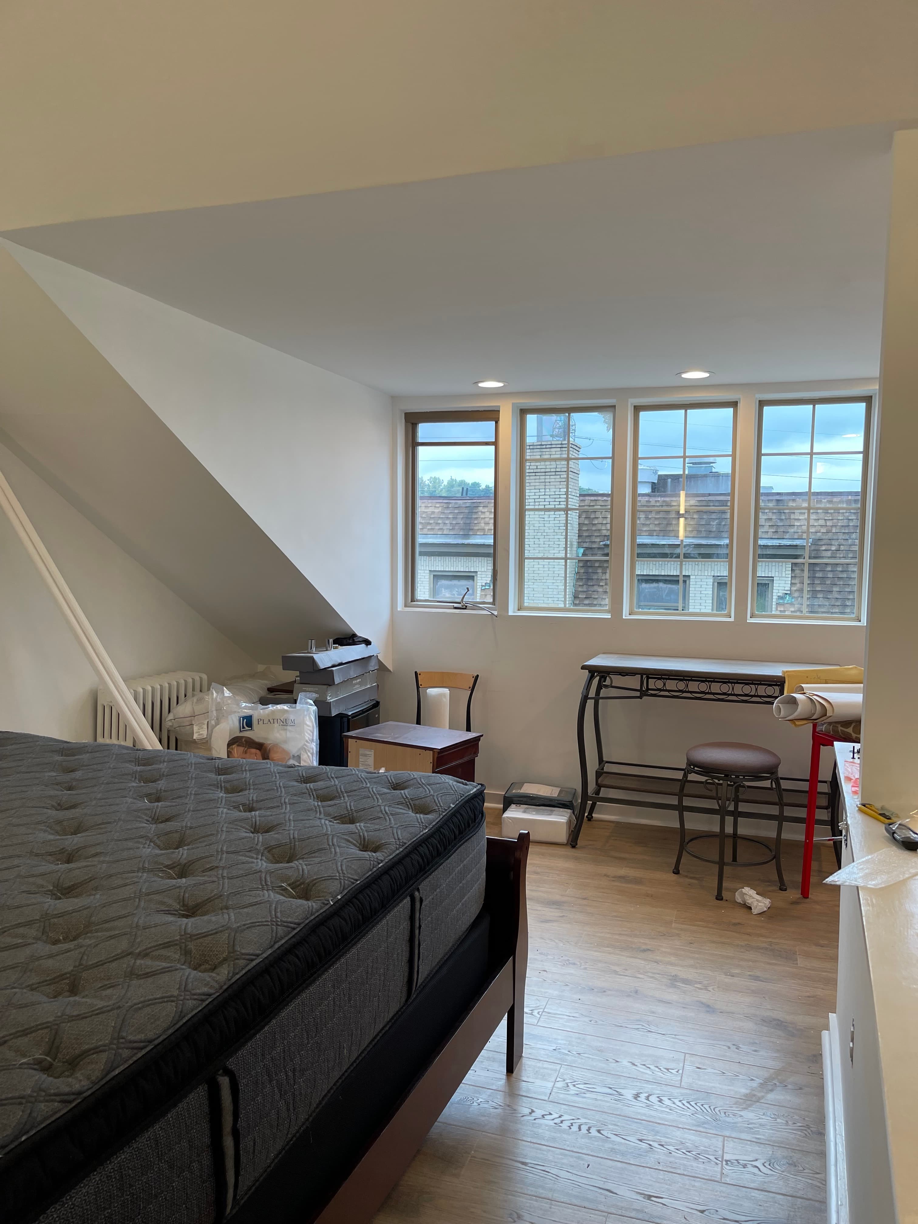 A loft-style bedroom with a dark gray bed, a sloped ceiling, and a desk with a stool near a window.