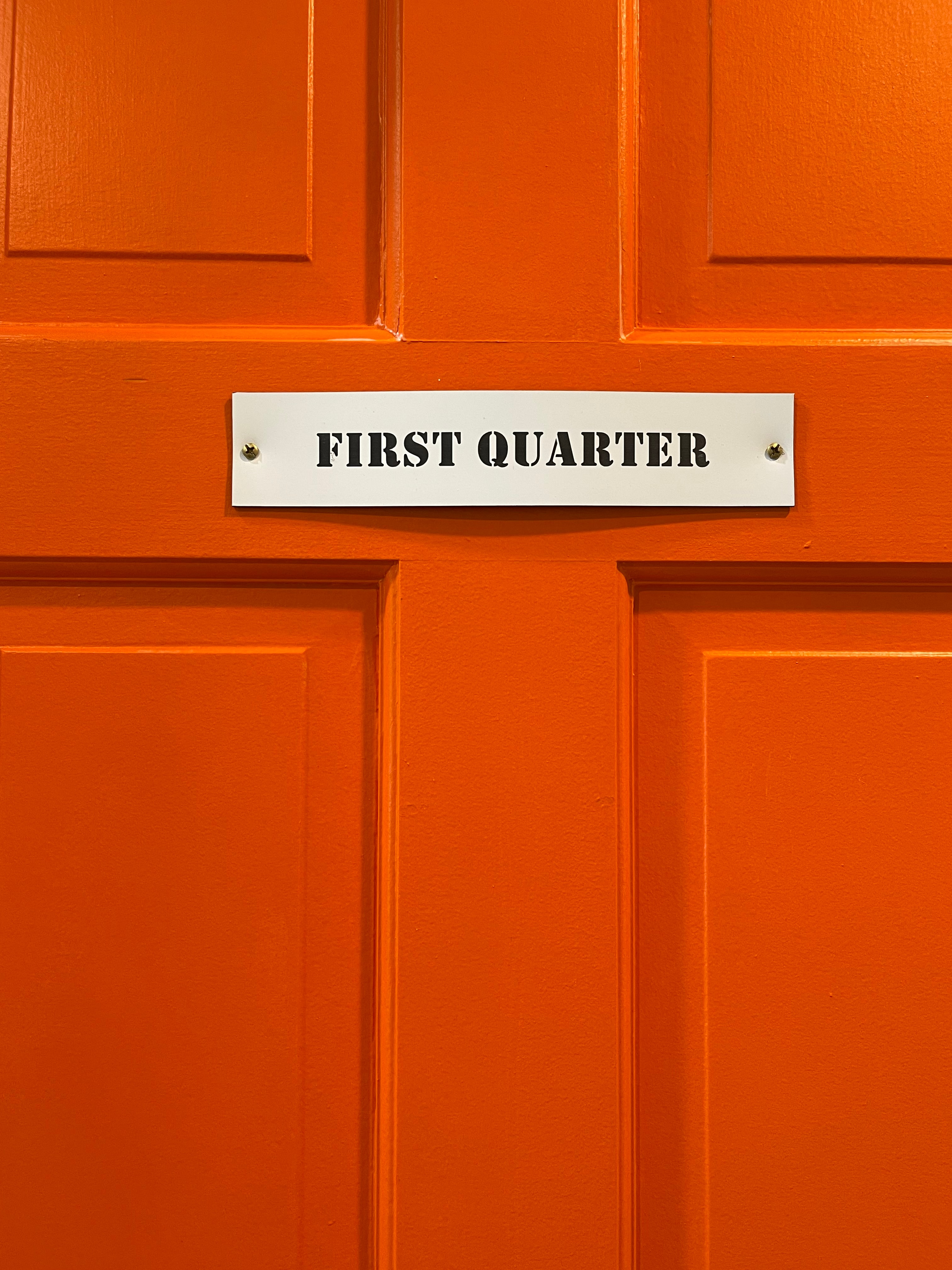 A rectangular white sign with black text that reads "FIRST QUARTER" attached to a vibrant orange paneled door.