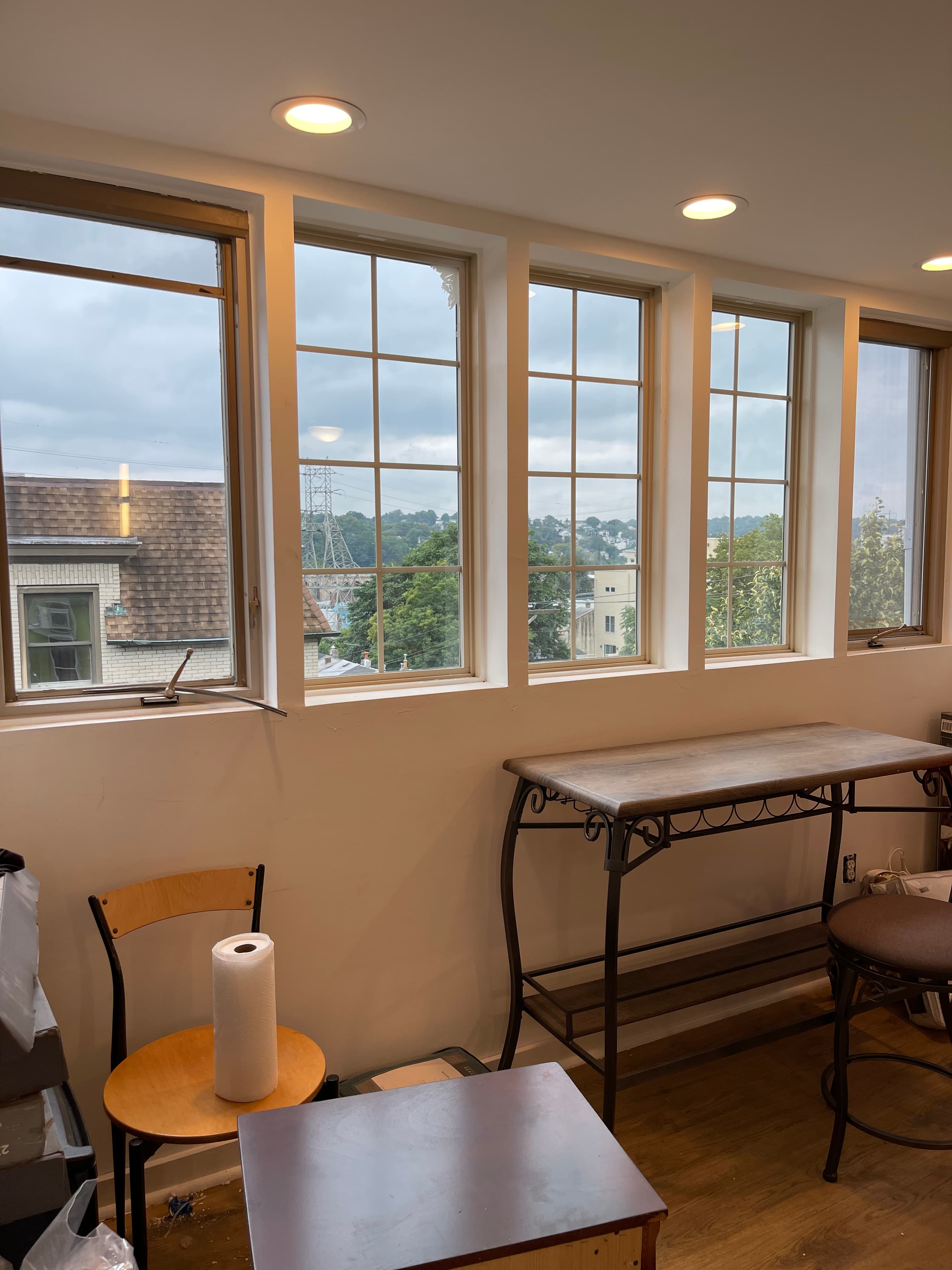 A room with four large windows offering a view of a cityscape. Two tables and a chair are visible in the foreground.