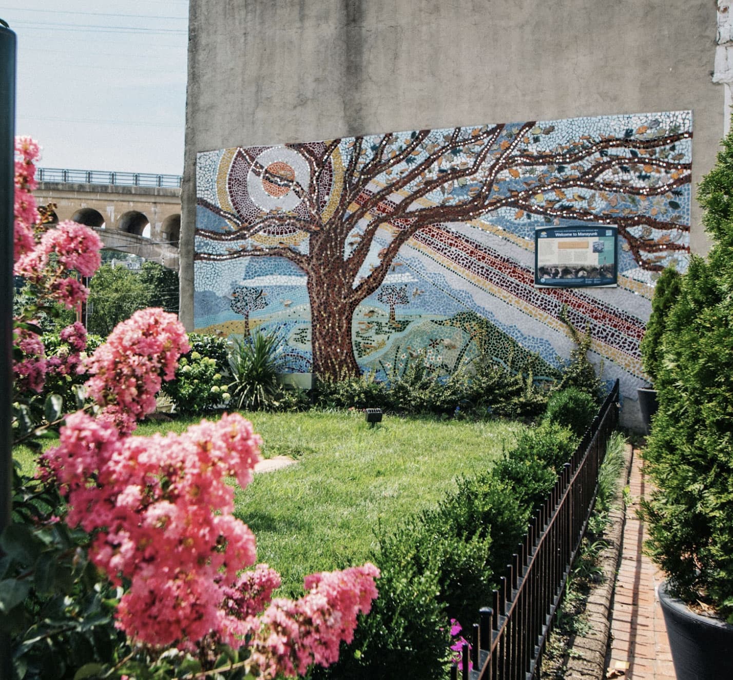 A colorful mosaic mural of a tree on a wall, surrounded by blooming flowers and greenery.
