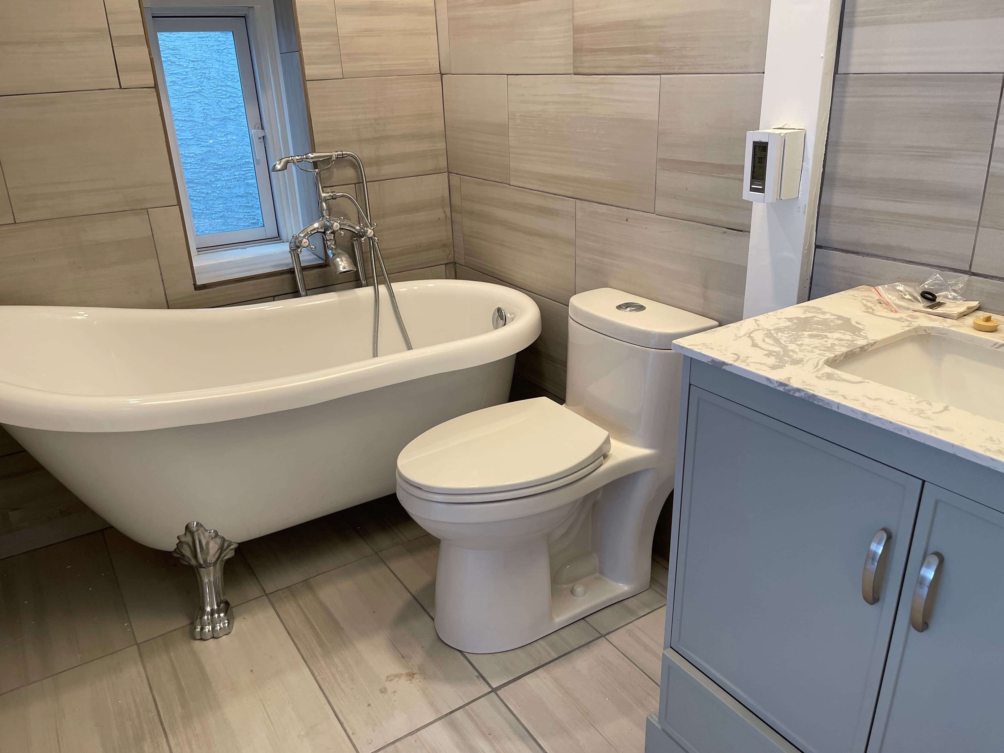 A modern bathroom with a freestanding soaking tub, a toilet, and a vanity with a sink. The walls are tiled with large, light-colored tiles.