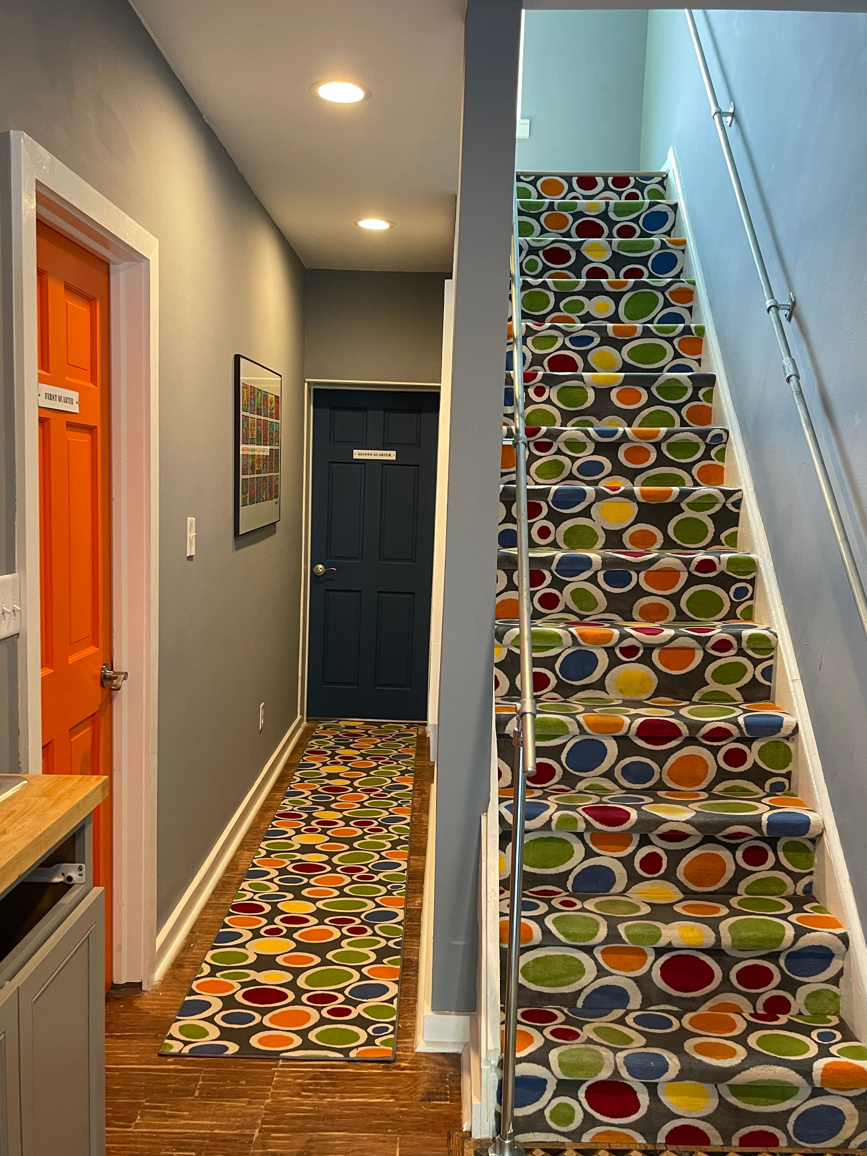 Long hallway with hardwood floors, leading to a staircase with a vibrant, multi-colored pattern. Two doors, one orange and one blue, are visible along the hallway.