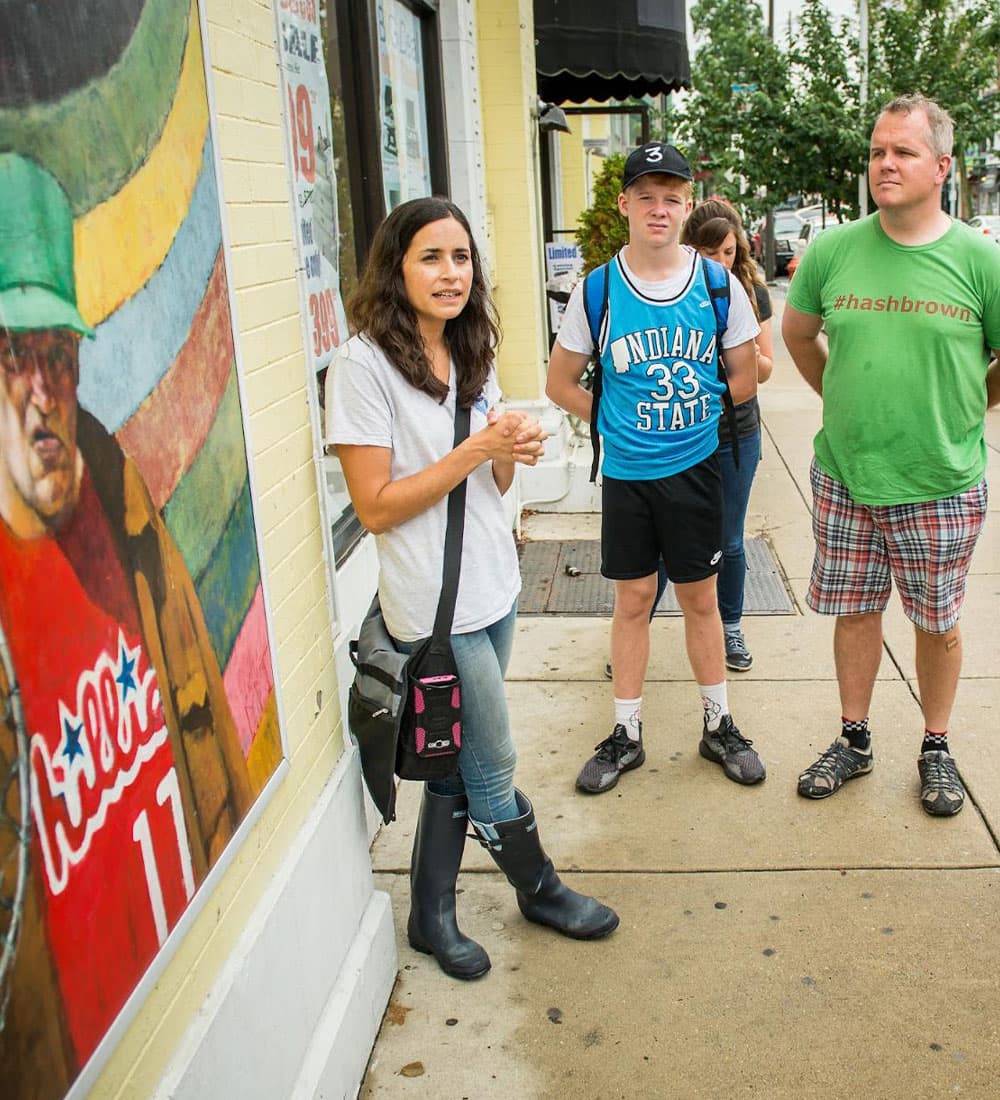 A group of people stands on a sidewalk near a colorful mural, with one woman speaking animatedly.