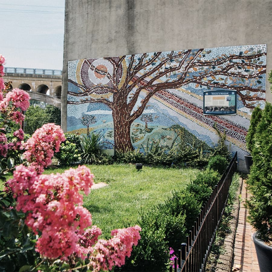 A vibrant mosaic mural of a tree, set against a grassy area with pink flowers and a nearby sign.