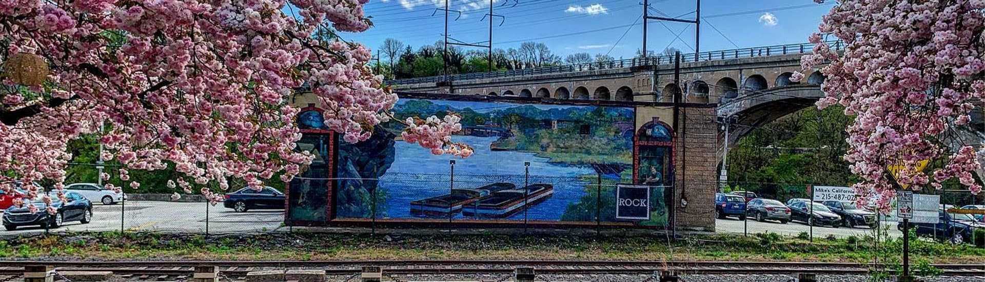 A vibrant mural depicting a river scene is framed by blooming cherry blossom trees and an arched bridge in the background.
