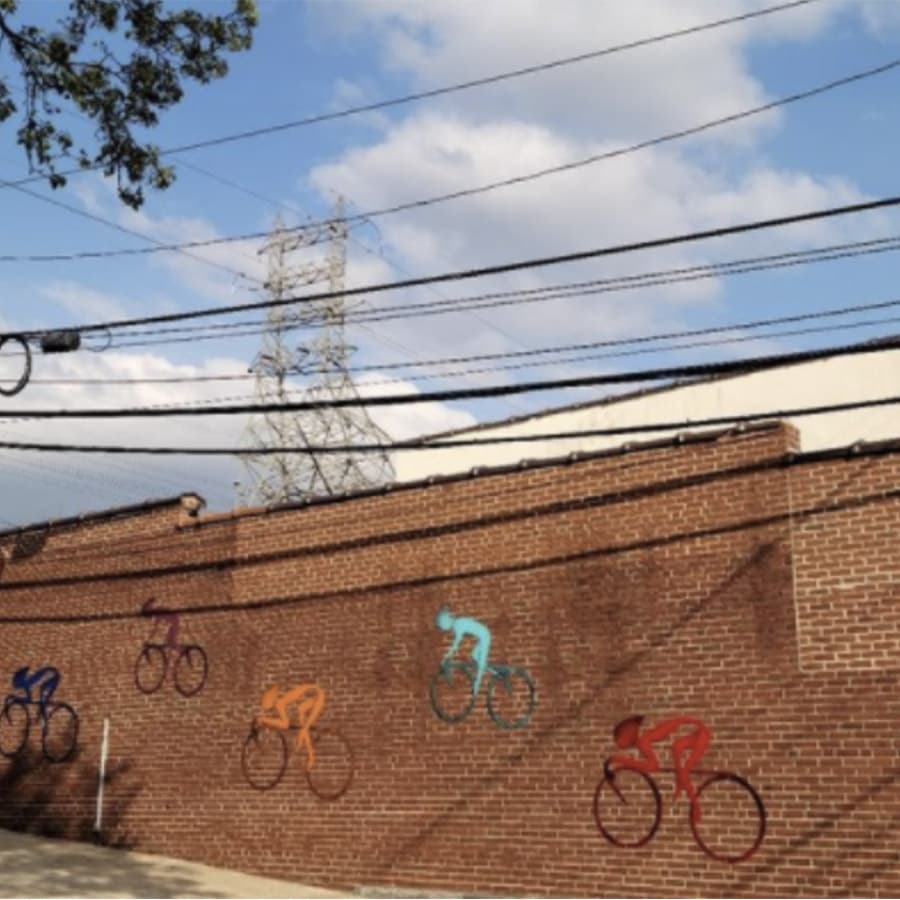Mural of colorful cyclists on a brick wall with electrical wires and a power tower in the background.