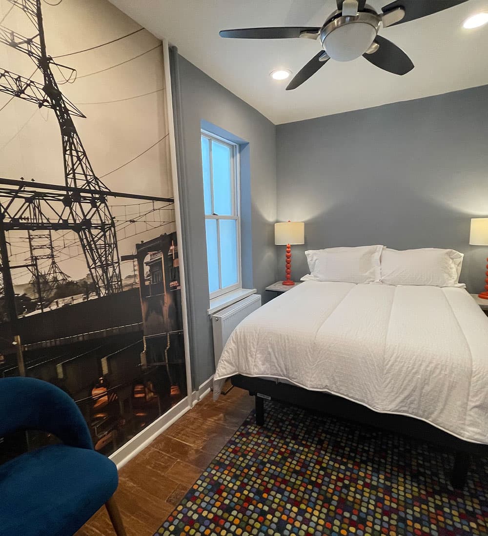 A cozy bedroom featuring a bed, colorful rug, modern lamps, and a mural of a power tower on the wall.