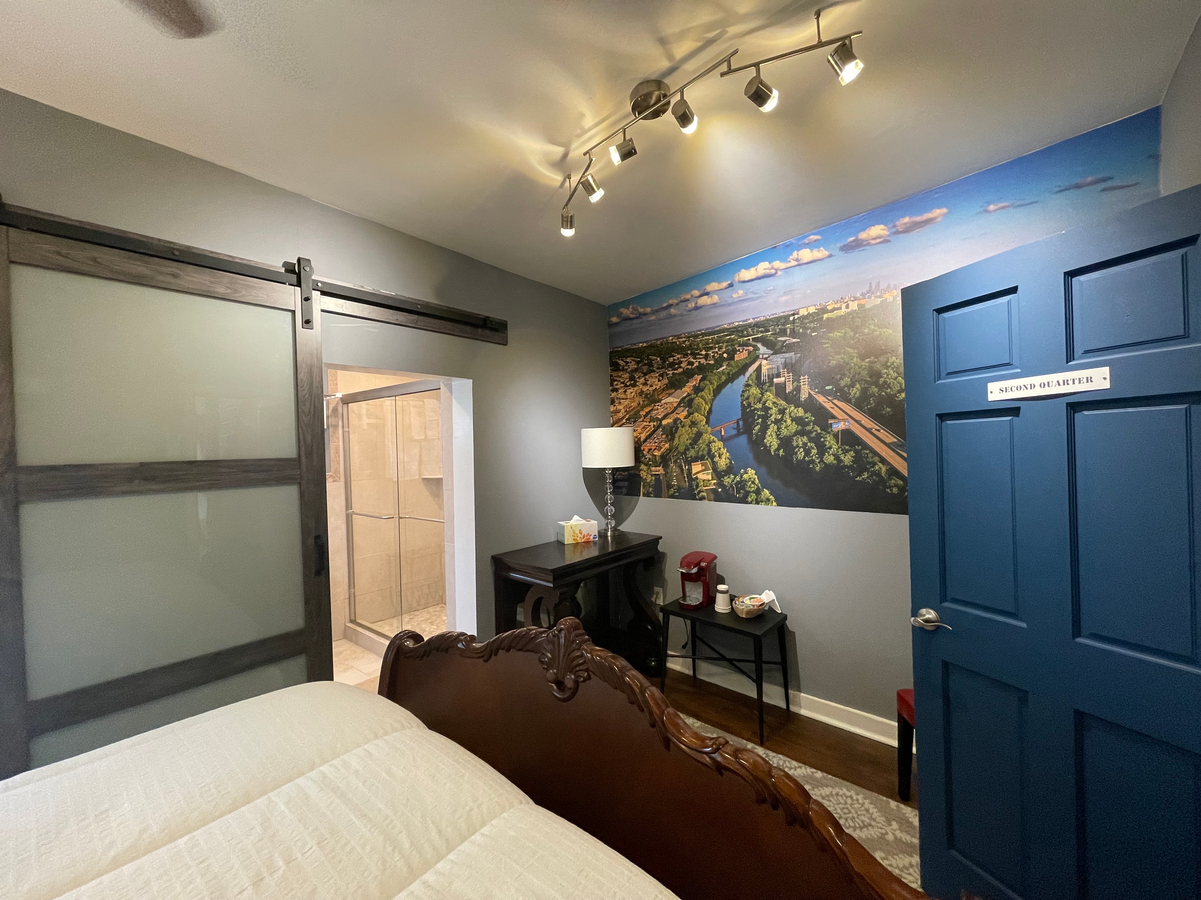 A modern bedroom with a large sliding barn door, a blue accent wall with a landscape mural, a dark blue door, and a glimpse of a bathroom through an open door.