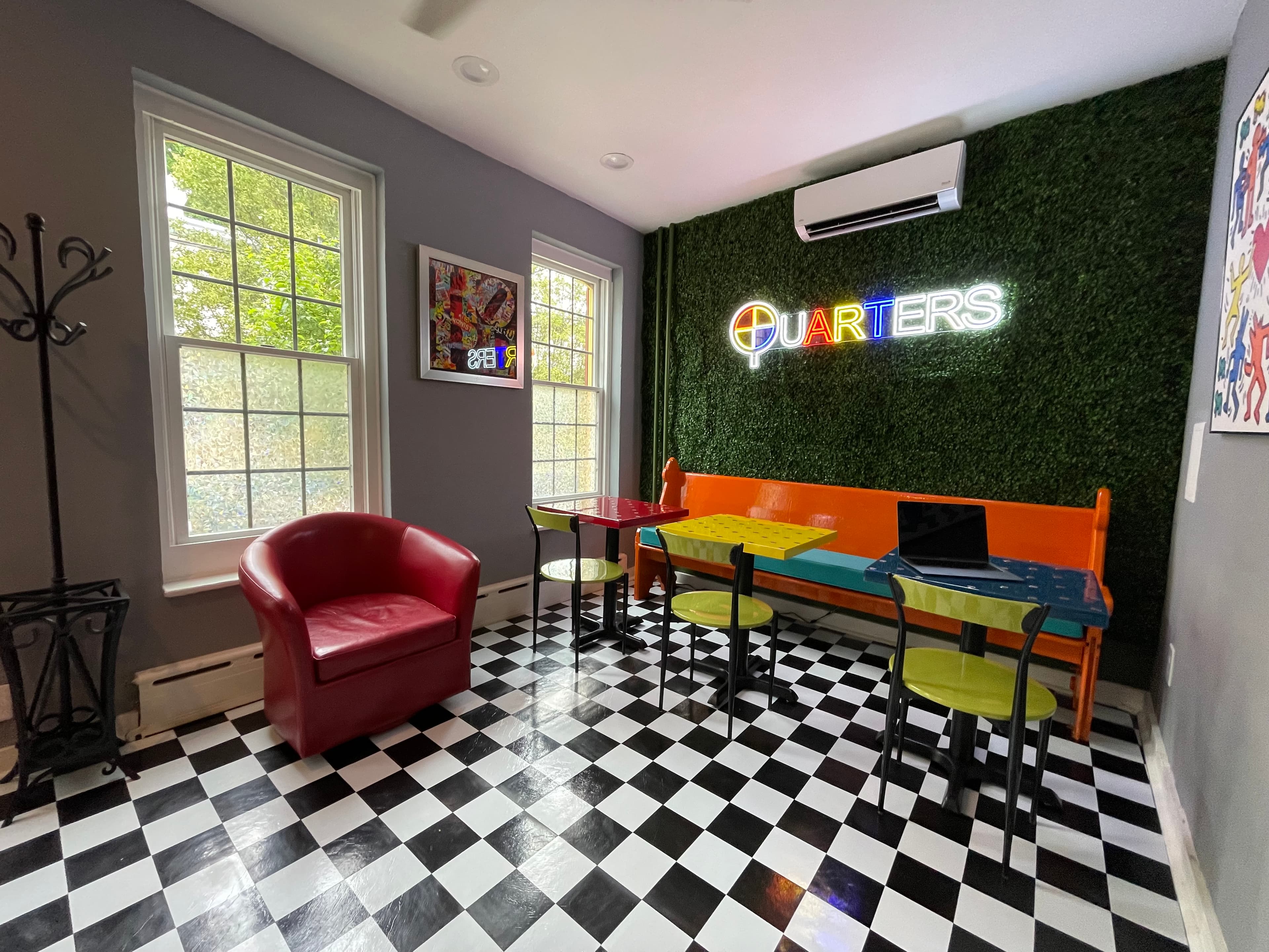 Dining area with black and white checkered floors, colorful tables and chairs, a red armchair, and a "Quarters" neon sign on a textured green wall.