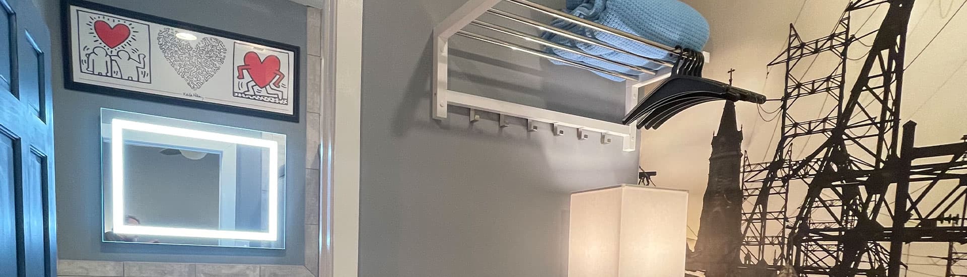 A corner of a room features a lighted mirror, a wall-mounted rack with a blue blanket, and artwork above.