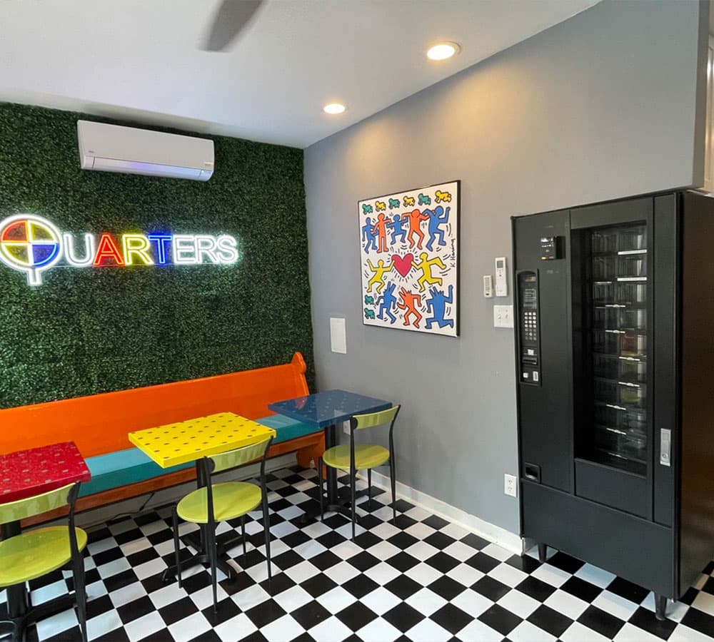 A colorful interior of a café featuring a neon sign, vibrant tables, and a vending machine.