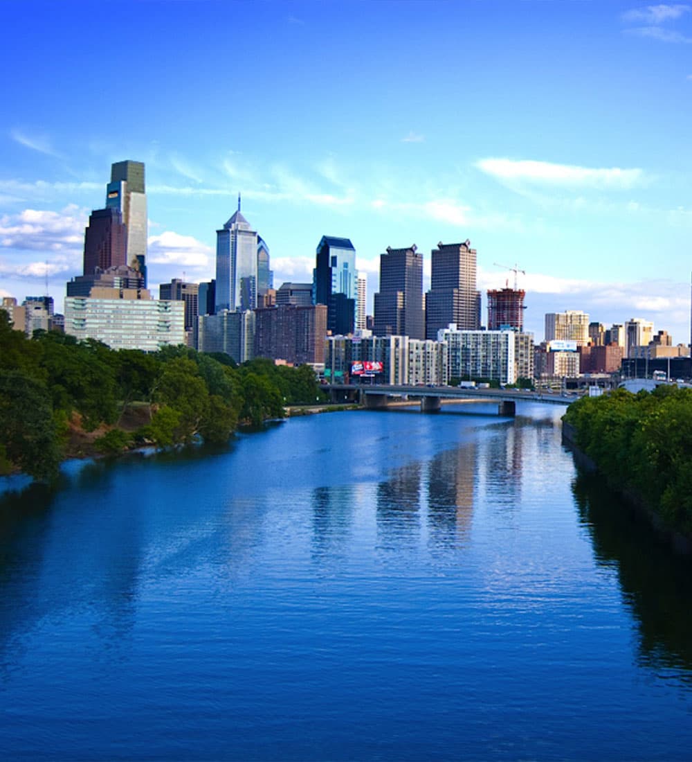 City skyline reflecting on a river under a clear blue sky.