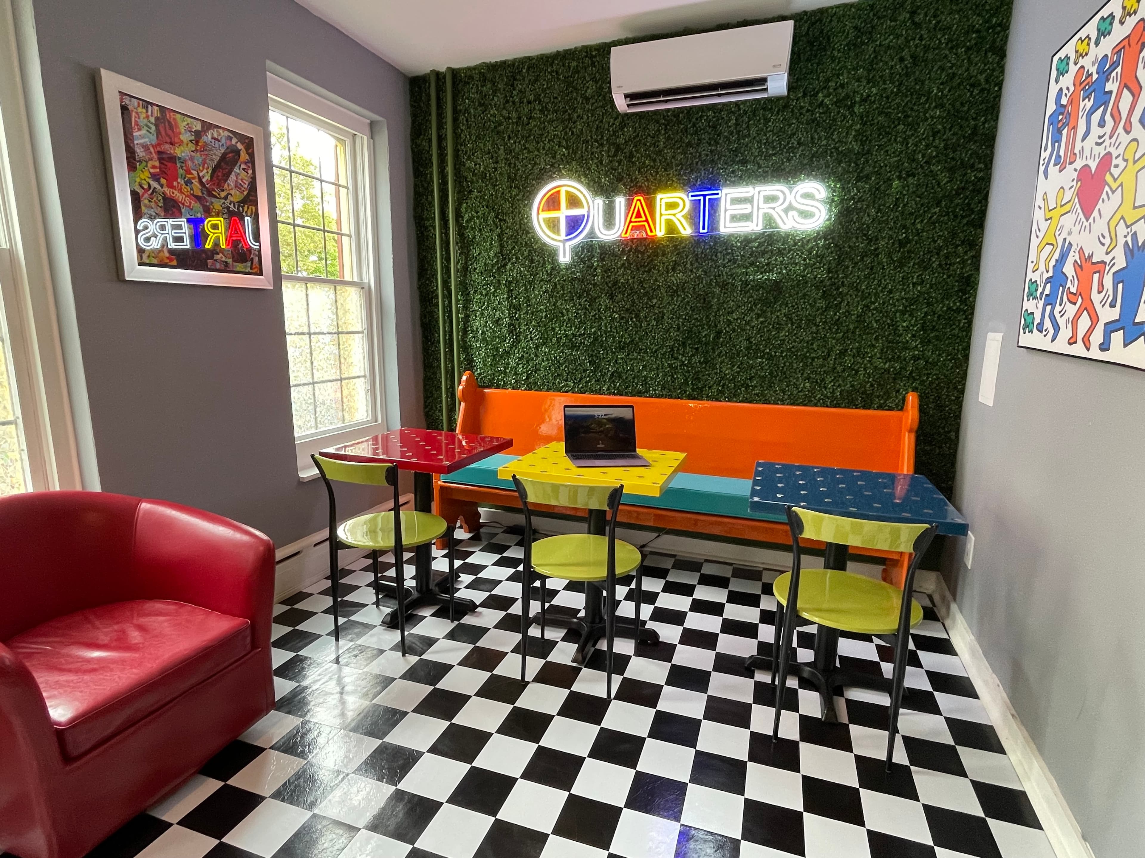Dining area with a "Quarters" neon sign on a green textured wall, featuring several tables, a red armchair, and artwork on the walls.