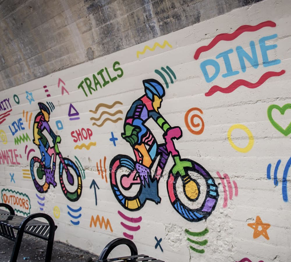 Colorful mural depicting two cyclists against playful, vibrant patterns and words like "TRAILS" and "DINE."