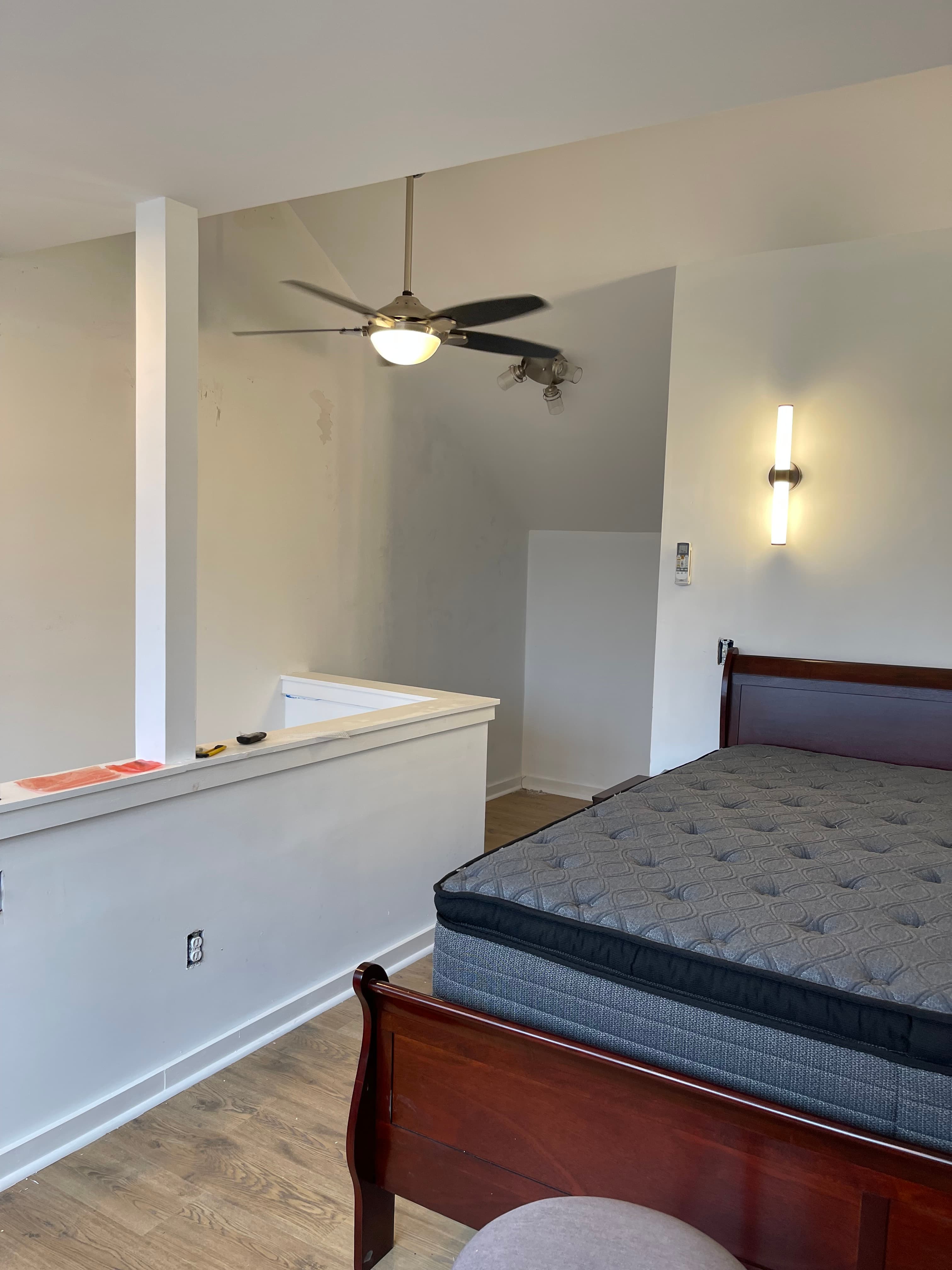 A loft-style bedroom with a dark wood bed frame, a gray mattress, and a white wall separating it from a lower level. A ceiling fan is visible above.