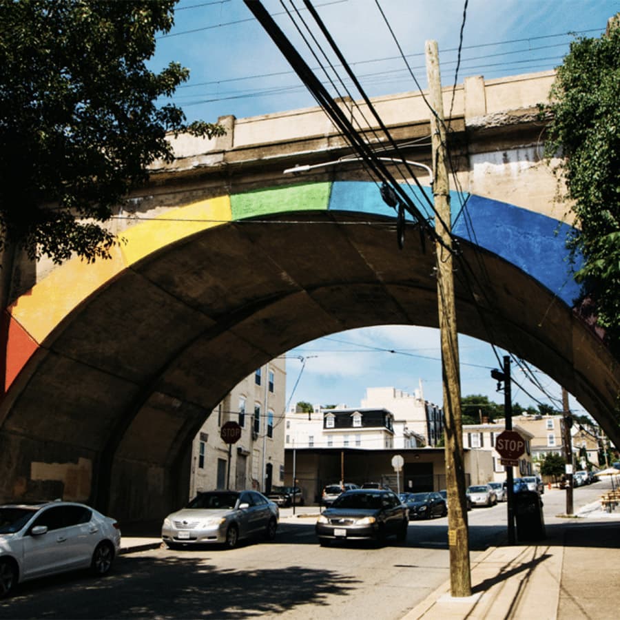 A colorful mural decorates an arching bridge over a busy street.